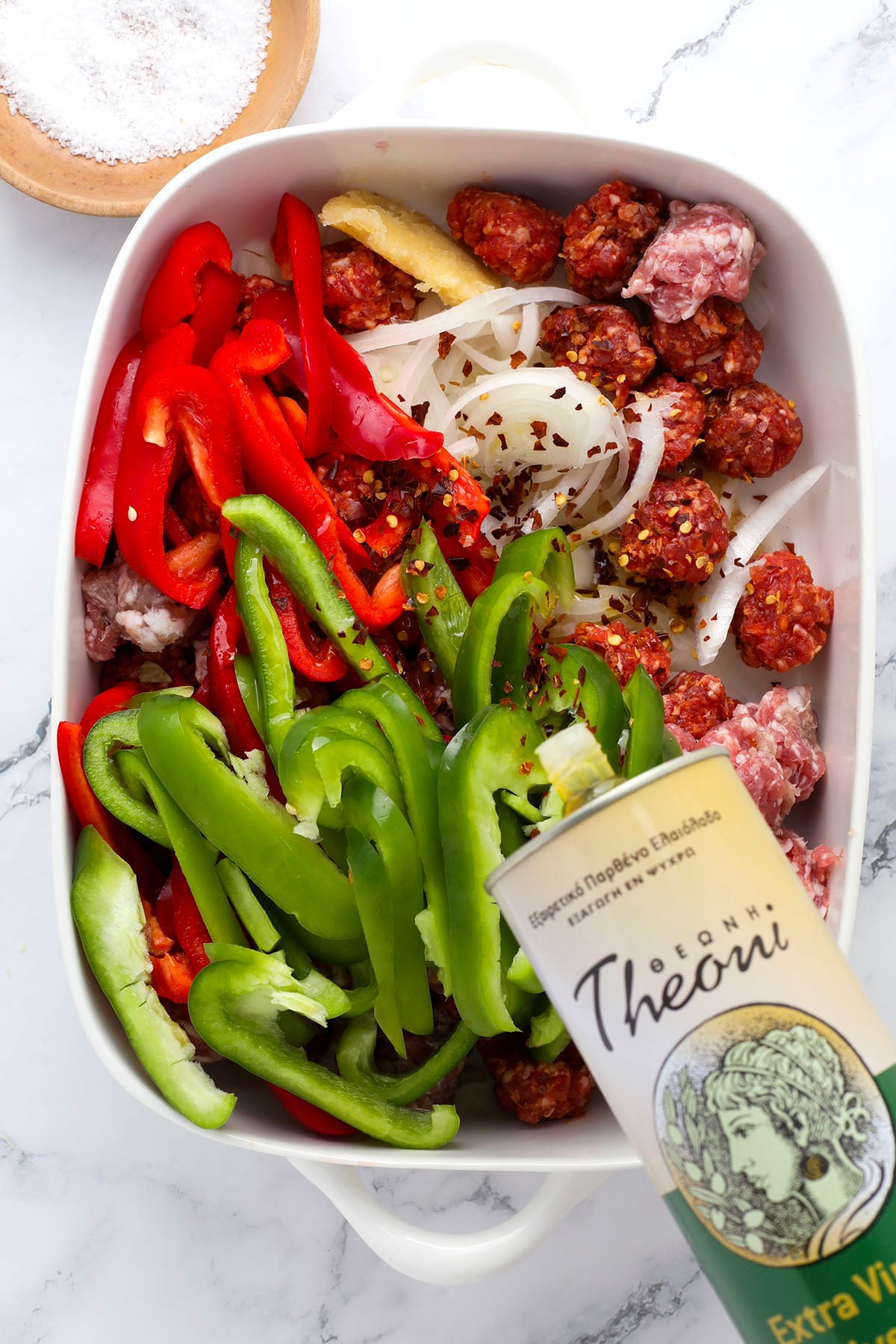 A white baking dish contains sliced red and green bell peppers, onions, and chunks of sausage. A hand pours extra virgin olive oil from a tin can onto the ingredients. A small bowl of salt sits nearby on a marble surface.