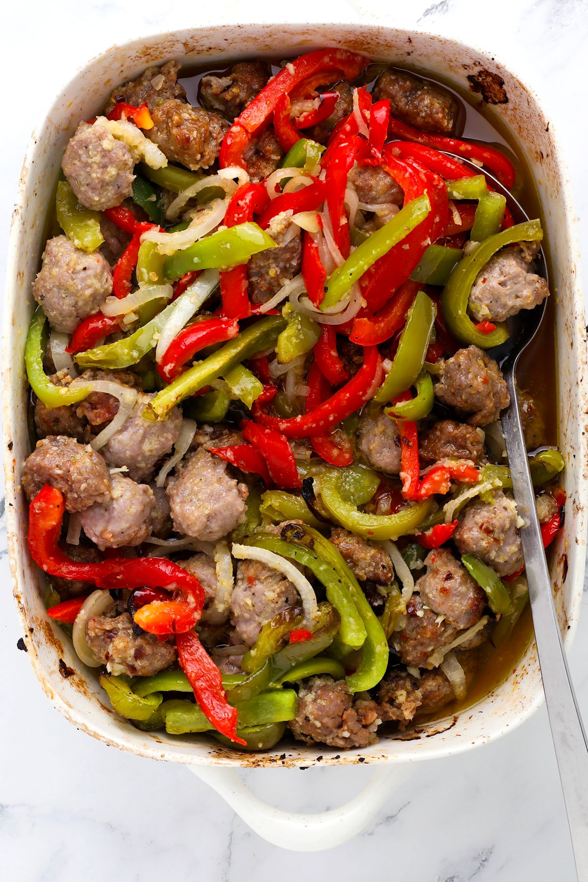 A baking dish filled with cooked sausage pieces, sliced red and green bell peppers, and onions, all mixed together with a light broth. A metal serving spoon rests inside the dish.