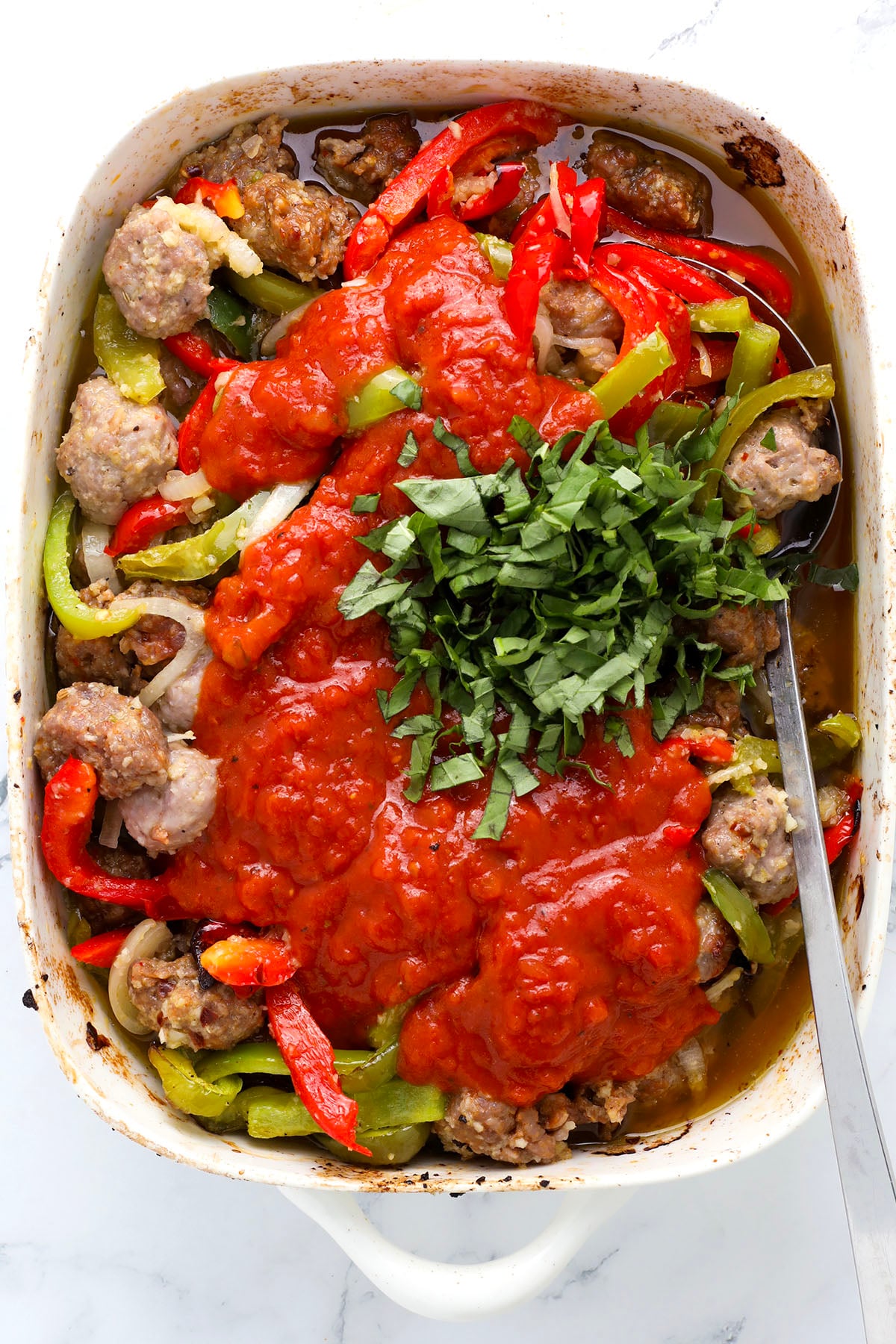 A white baking dish filled with cooked sausage pieces, sliced red and green bell peppers, onions, topped with tomato sauce and fresh chopped basil, with a serving spoon on the side.