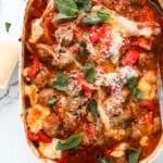 A baked dish of meatballs with melted cheese, tomato sauce, red bell peppers, and fresh basil leaves, served in a white casserole dish on a marble surface.