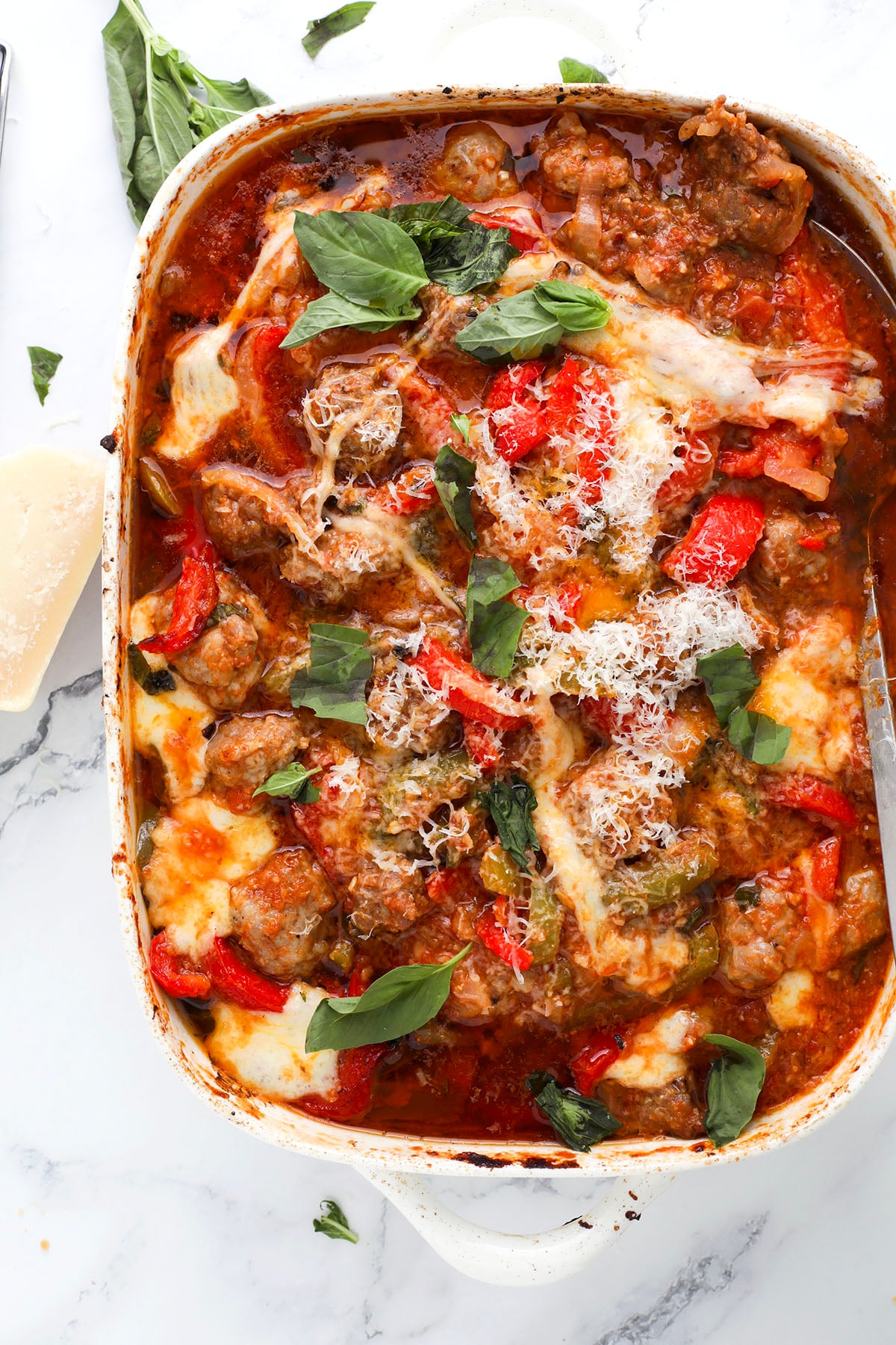 A baked dish of meatballs with melted cheese, tomato sauce, red bell peppers, and fresh basil leaves, served in a white casserole dish on a marble surface.