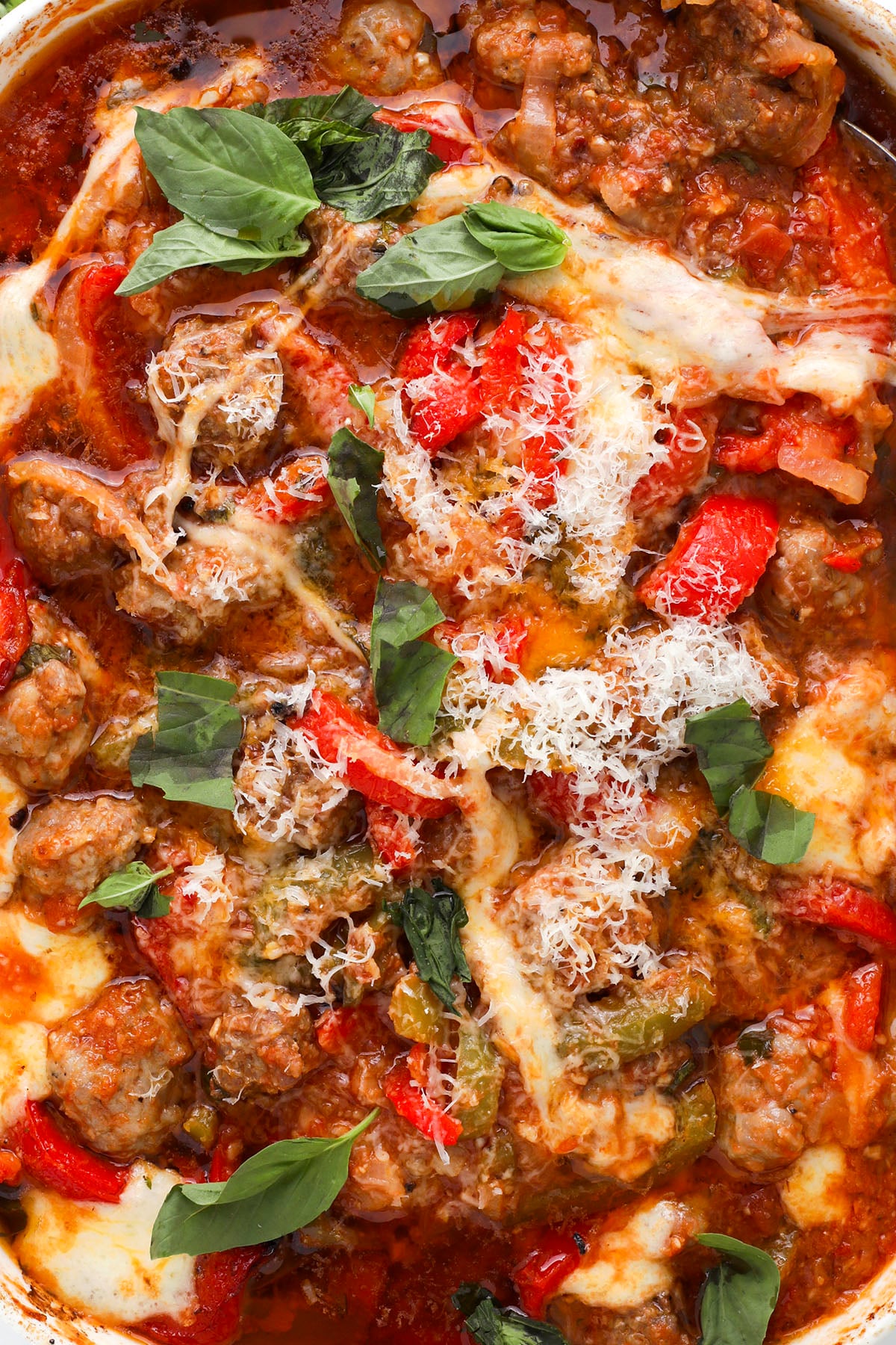 A close-up of a baked Italian sausage and pepper dish topped with melted cheese, grated Parmesan, fresh basil leaves, and slices of red and green bell peppers in a rich tomato sauce.
