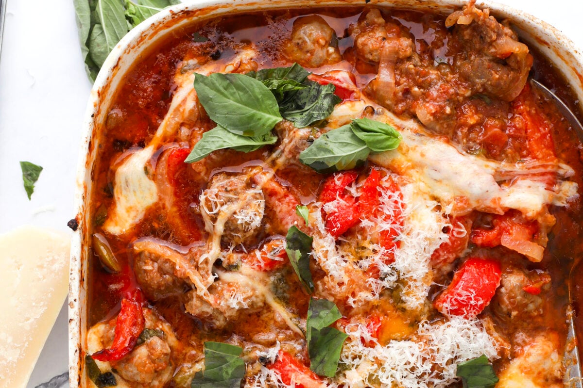 A close-up of a baked Italian dish with meatballs, melted cheese, tomato sauce, red peppers, grated parmesan, and fresh basil leaves on top in a white baking dish.