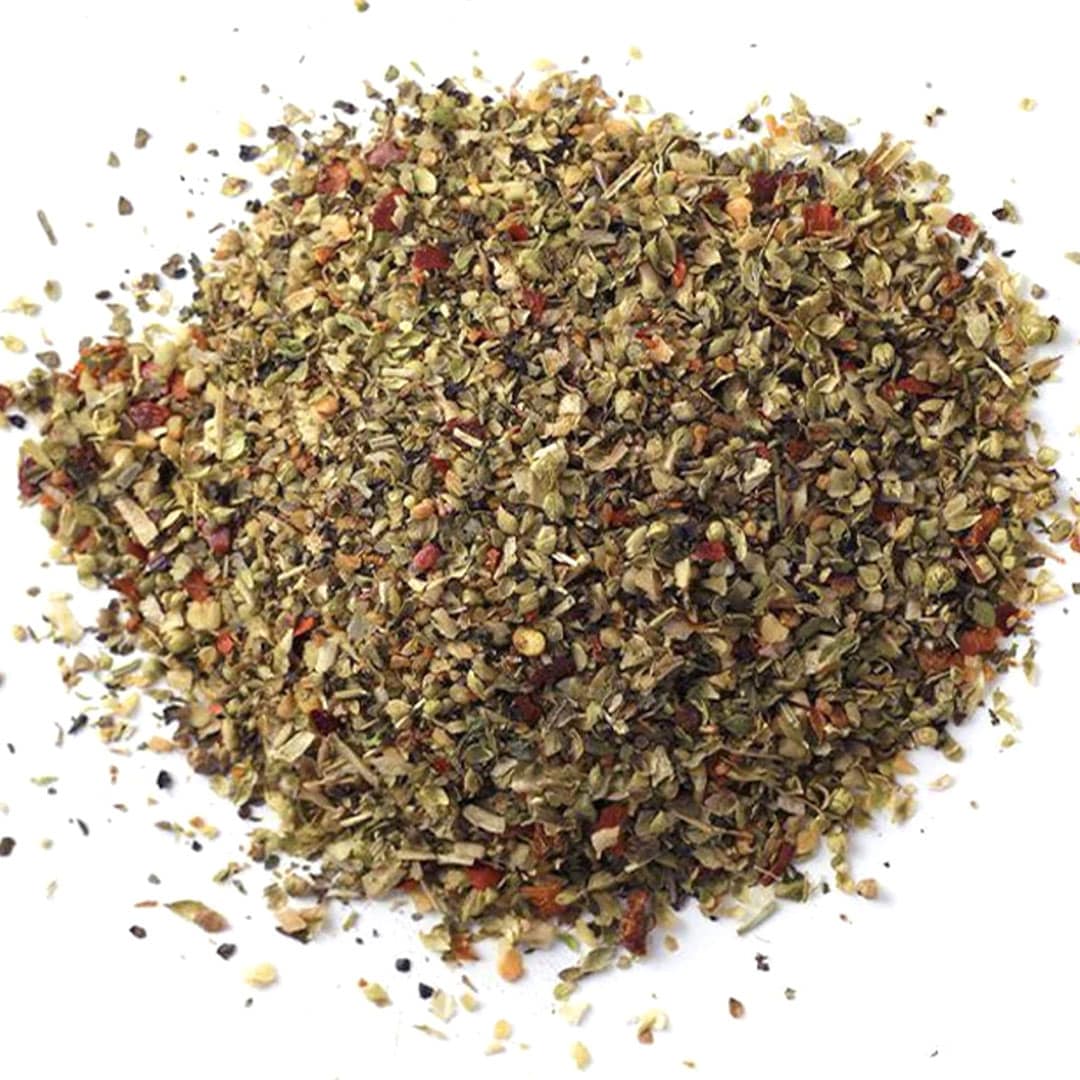 A pile of mixed dried Italian herbs and spices, featuring green, red, and brown flakes, scattered on a white background.