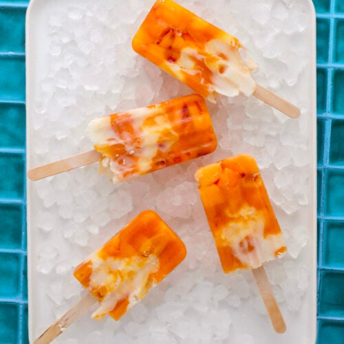 Four orange and white swirled popsicles are arranged on a white tray filled with ice cubes, set on a bright blue tiled surface.