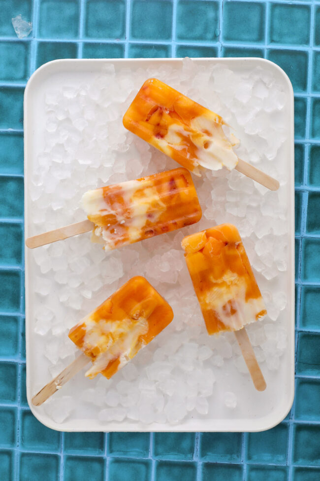 Four orange and white swirled popsicles are arranged on a white tray filled with ice cubes, set on a bright blue tiled surface.