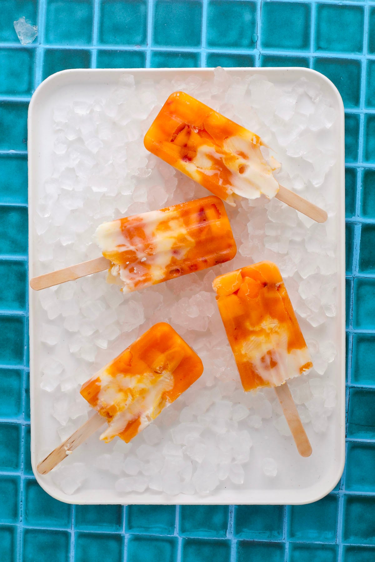 Four orange and white swirled popsicles are arranged on a white tray filled with ice cubes, set on a bright blue tiled surface.