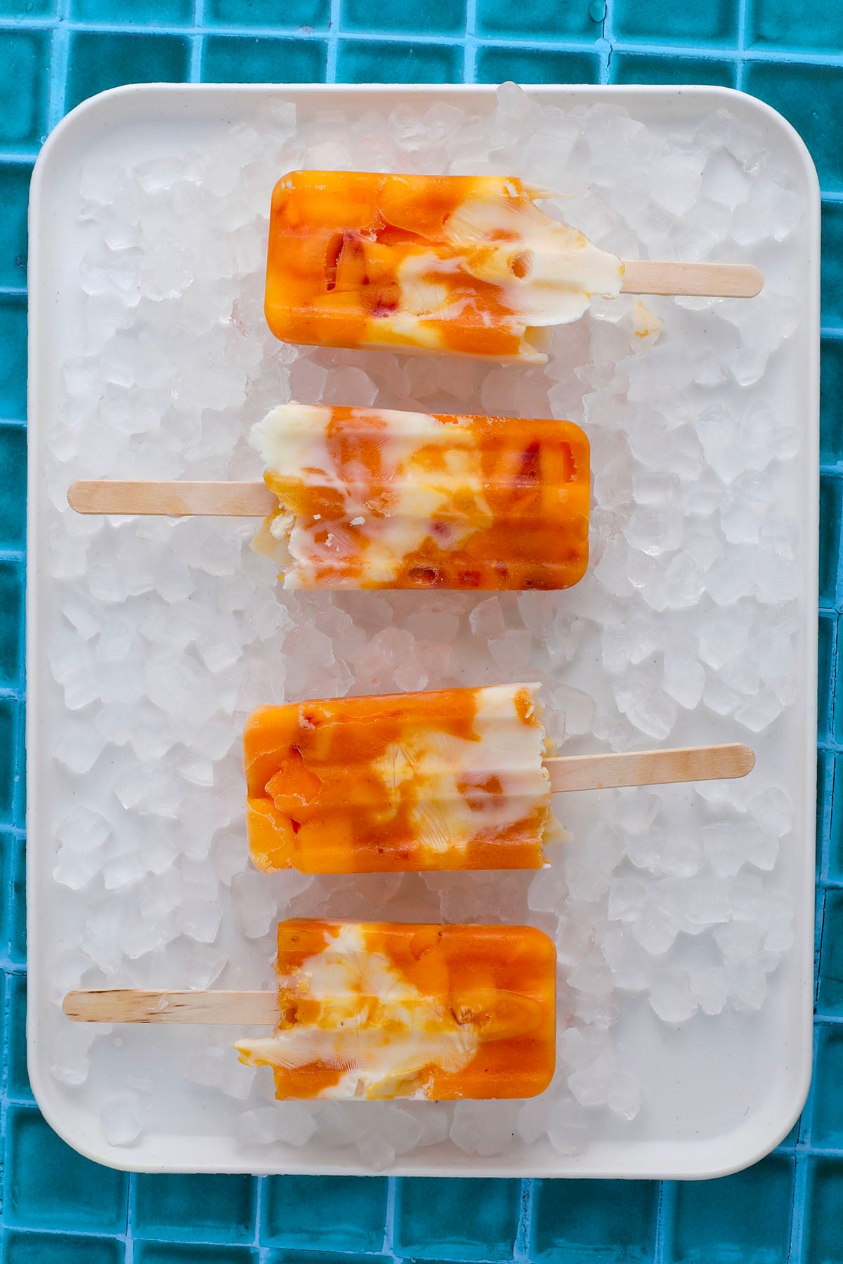 Four orange and white popsicles with a marbled pattern are arranged on a white tray filled with ice cubes, set on a blue tiled surface.