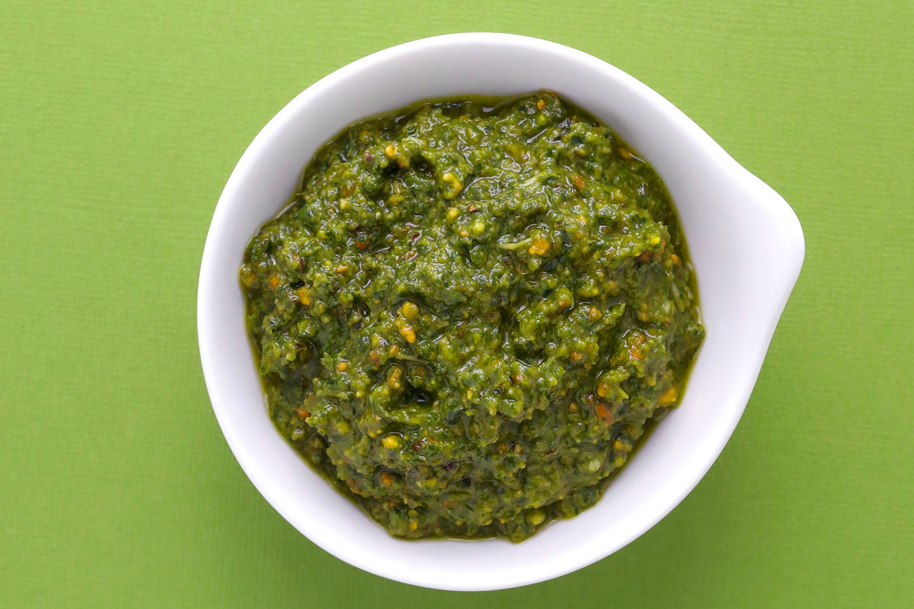 A white bowl filled with green pesto sauce sits on a green surface. The pesto appears thick, textured, and speckled with small bits of herbs and nuts.