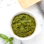 A bowl of green pesto sauce sits on a white marble surface next to a sprig of fresh basil, a wedge of Parmesan cheese, and a metal grater.