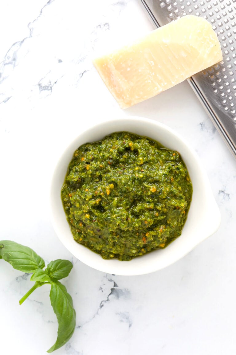 A bowl of green pesto sauce sits on a white marble surface next to a sprig of fresh basil, a wedge of Parmesan cheese, and a metal grater.