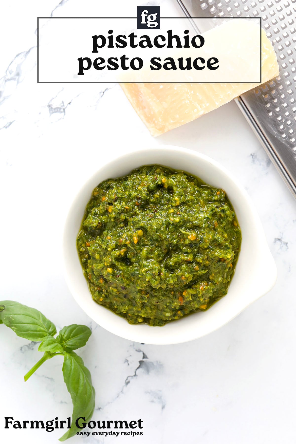 A small white bowl filled with vibrant pesto with pistachio nuts sits on a marble surface next to a wedge of Parmesan, a grater, and fresh basil. Text reads recipe for pistachio pesto.