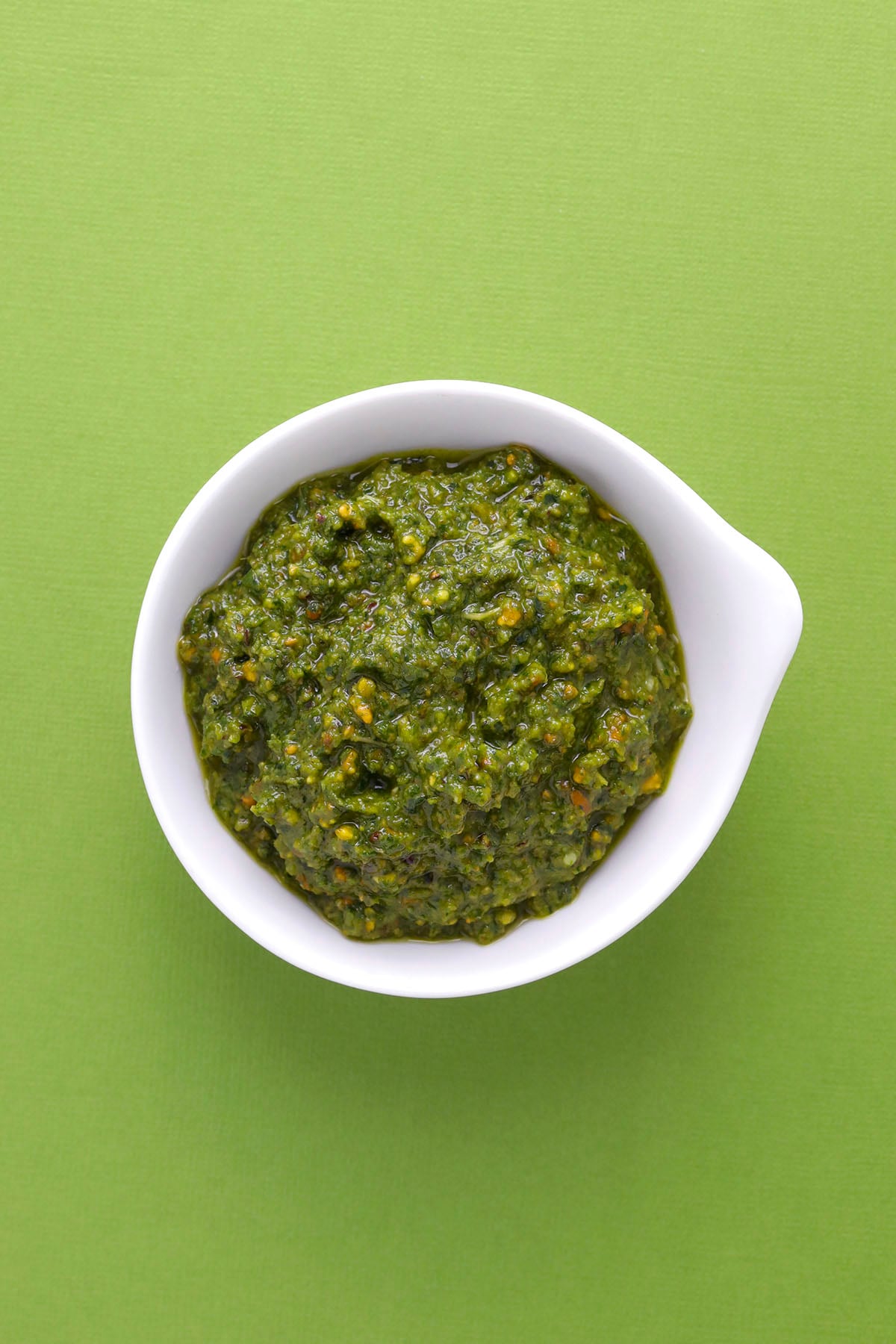 A small white bowl filled with vibrant green pistachio pesto sauce, placed on a solid light green background.