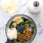 A food processor holds fresh basil, pistachios, Parmesan cheese, and a wedge of cheese—perfect for making pesto with pistachio. Nearby sit a bowl of shredded cheese and a white bottle, all arranged on a marble counter.