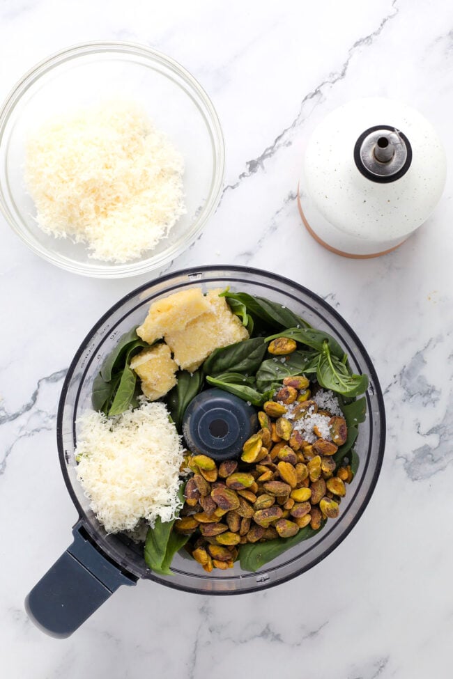 A food processor holds fresh basil, pistachios, Parmesan cheese, and a wedge of cheese—perfect for making pesto with pistachio. Nearby sit a bowl of shredded cheese and a white bottle, all arranged on a marble counter.