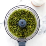 A food processor bowl filled with freshly blended pesto with pistachio nuts sits on a white marble surface next to a white oil dispenser bottle.