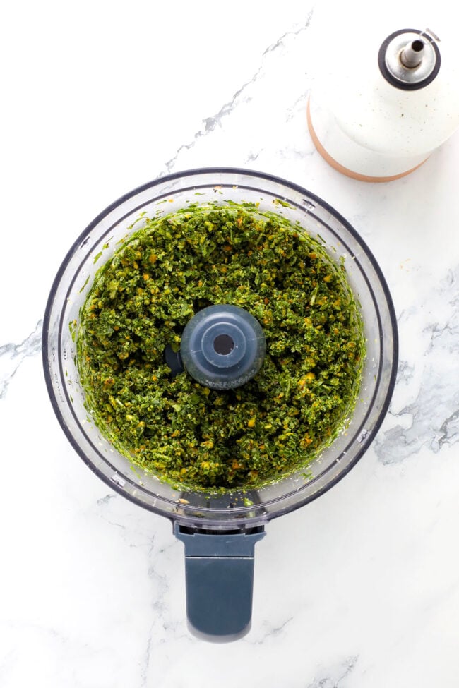 A food processor bowl filled with freshly blended pesto with pistachio nuts sits on a white marble surface next to a white oil dispenser bottle.