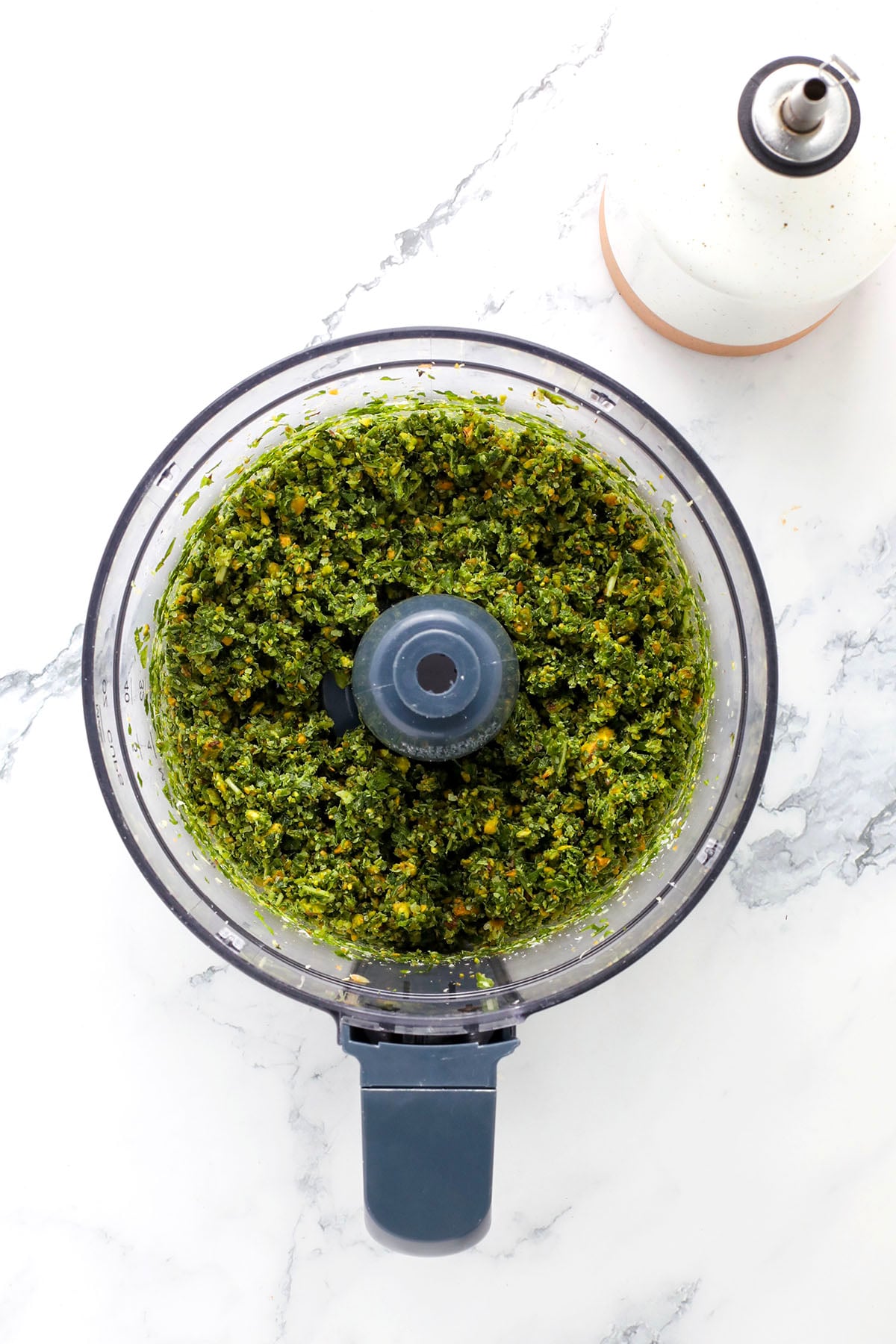 A food processor holds fresh basil, pistachios, Parmesan cheese, and a wedge of cheese—perfect for making pesto with pistachio. Nearby sit a bowl of shredded cheese and a white bottle, all arranged on a marble counter.