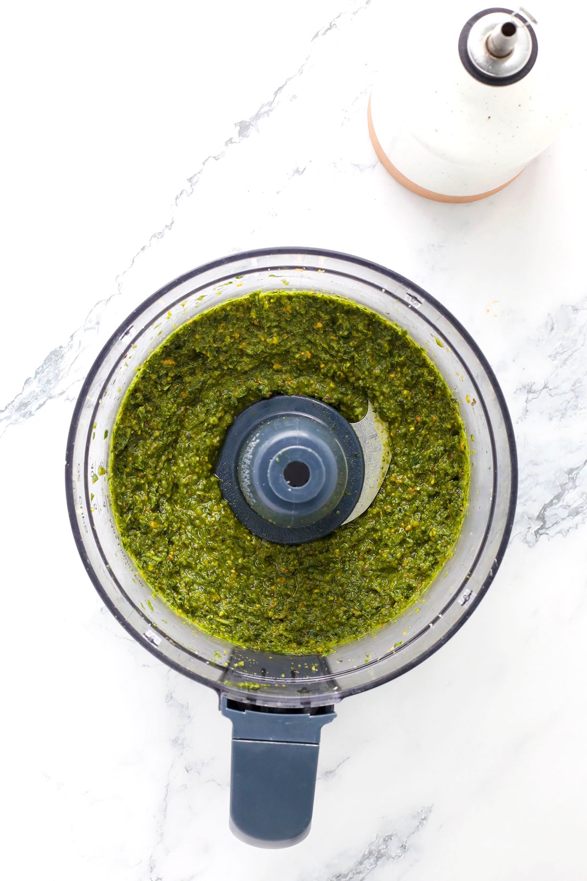 A food processor containing freshly blended pistachio pesto sauce sits on a white marble countertop, with an olive oil dispenser nearby.