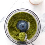 A hand holds a spoon of vibrant pesto with pistachio from a food processor on a marble countertop, with a bottle of oil visible in the corner.