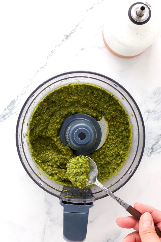 A hand holds a spoon of vibrant pesto with pistachio from a food processor on a marble countertop, with a bottle of oil visible in the corner.