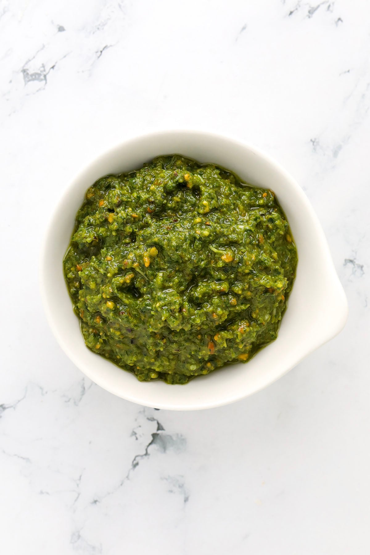 A small white bowl filled with vibrant pistachio pesto sauce, placed on a white marble surface, showcases a delicious recipe for pistachio pesto.