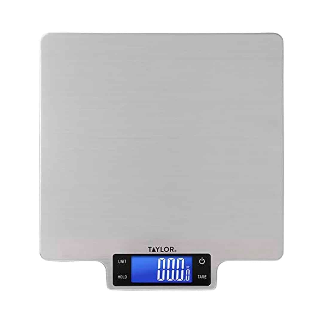 A digital kitchen scale with a stainless steel surface displays 1000 g on a blue backlit LCD screen, making it the perfect companion for following the ultimate homemade bread guide. The scale has buttons labeled unit, hold, and tare, and the brand name Taylor appears below the display.