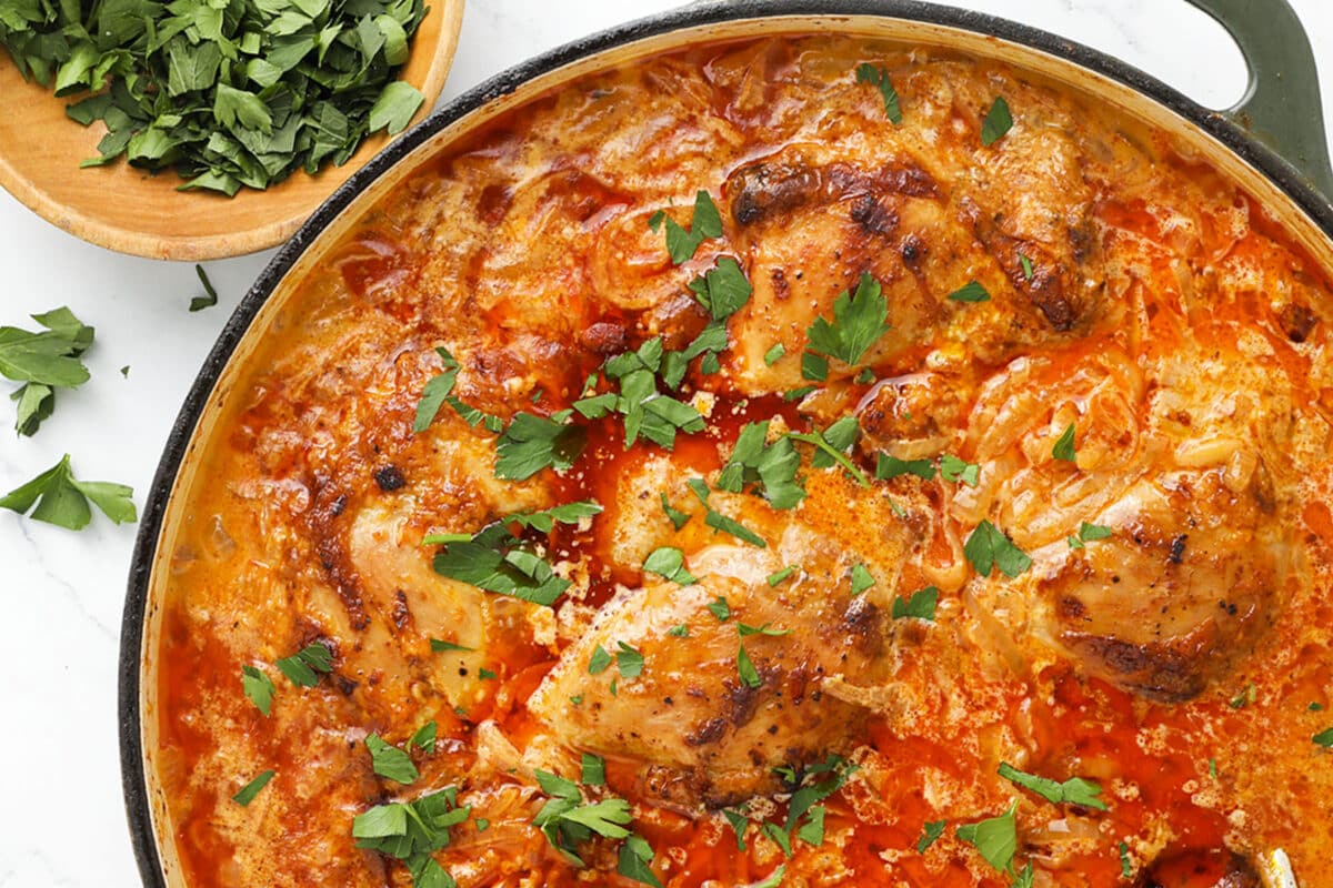Chicken thighs simmered in a creamy, tomato-based sauce topped with fresh chopped parsley in a skillet, with a bowl of extra parsley on the side.