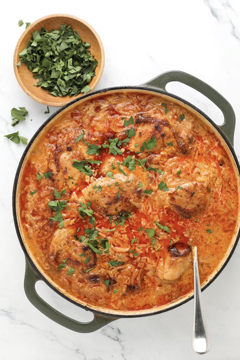 A pot of creamy tomato chicken and rice garnished with chopped parsley, with a spoon inside. A small wooden bowl of fresh chopped parsley sits nearby on a white marble surface.