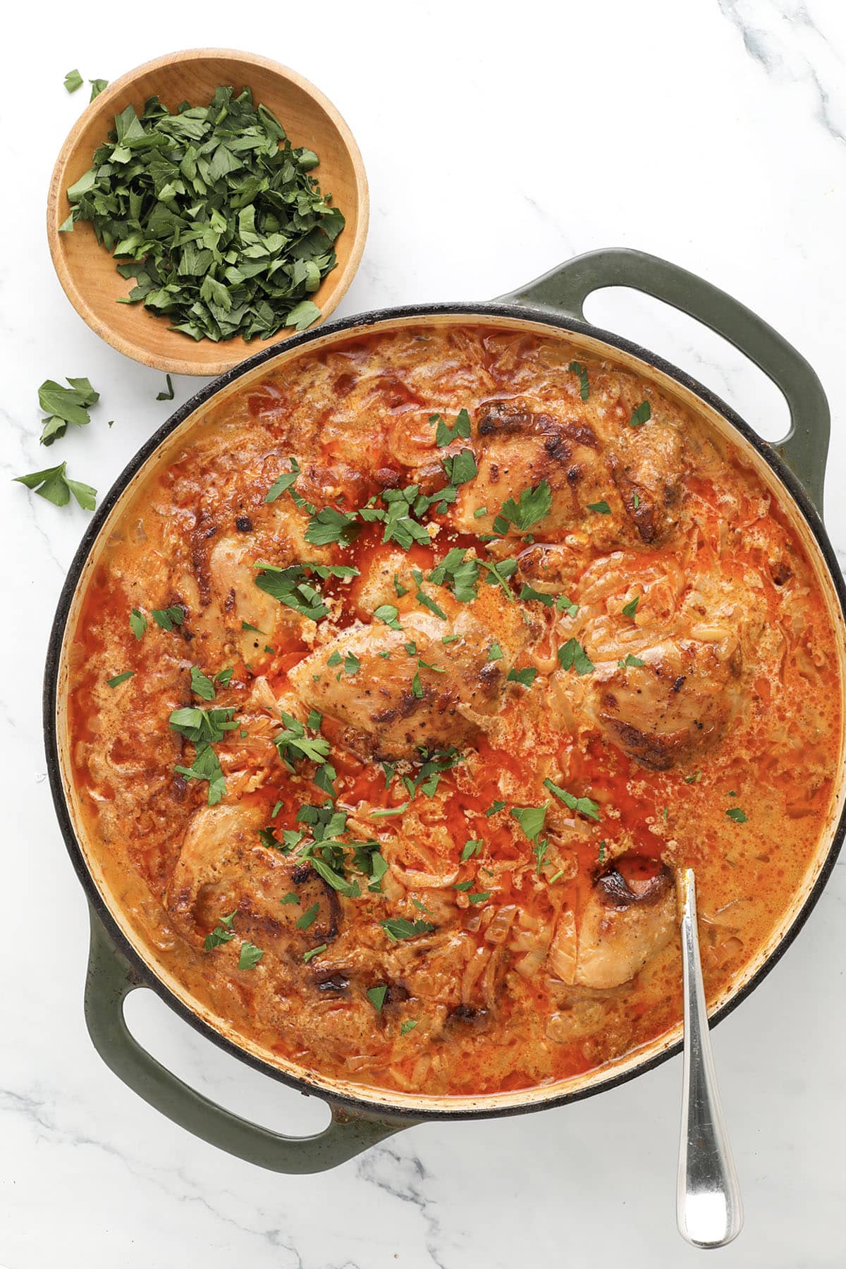 A pot of creamy tomato chicken and rice garnished with chopped parsley, with a spoon inside. A small wooden bowl of fresh chopped parsley sits nearby on a white marble surface.