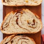 Three slices of cinnamon swirl bread with raisins are arranged on a wooden cutting board, showing their spiral pattern and soft texture.