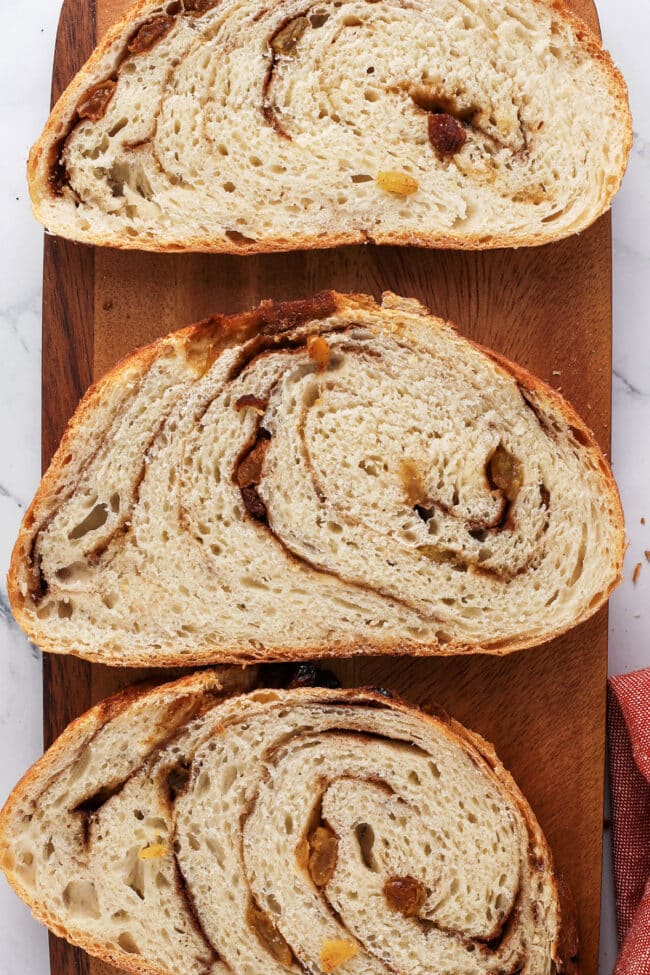 Three slices of cinnamon swirl bread with raisins are arranged on a wooden cutting board, showing their spiral pattern and soft texture.
