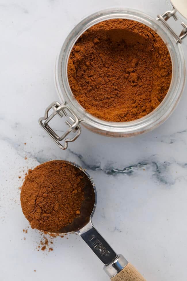 An open glass jar of ground cinnamon with a metal clasp sits on a white surface. Next to it lies a metal tablespoon filled with cinnamon, with some powder spilled beside it.