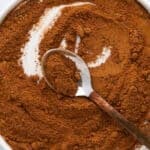 A round white dish filled with ground brown spice, possibly cinnamon, with a metal spoon resting inside, partially buried in the powder.