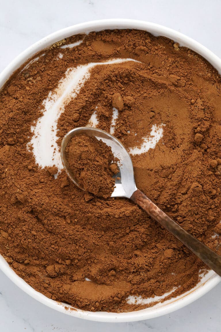 A round white dish filled with ground brown spice, possibly cinnamon, with a metal spoon resting inside, partially buried in the powder.