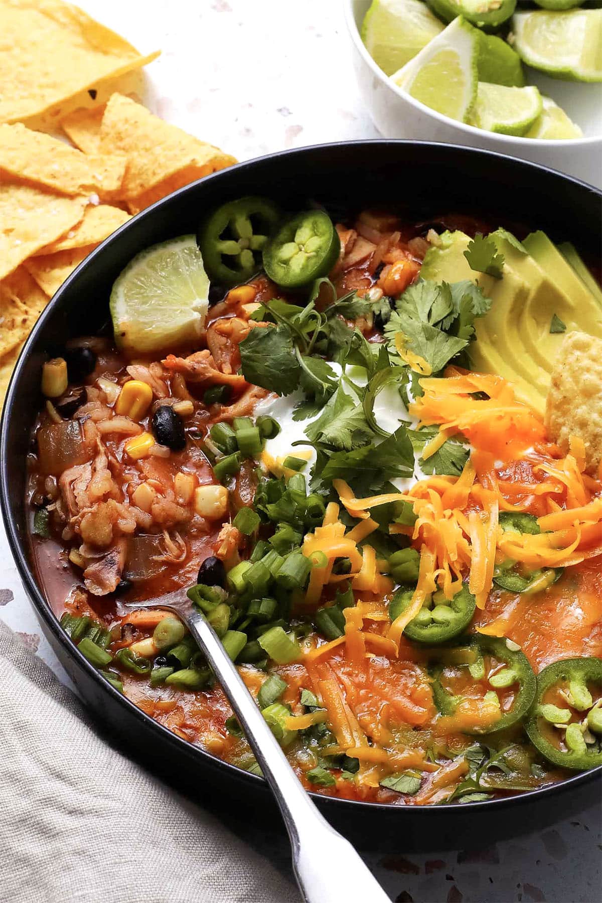 A bowl of rotisserie chicken tortilla soup topped with avocado, shredded cheese, jalapeño slices, cilantro, green onions, a lime wedge, and sour cream, with tortilla chips and a bowl of lime wedges on the side.