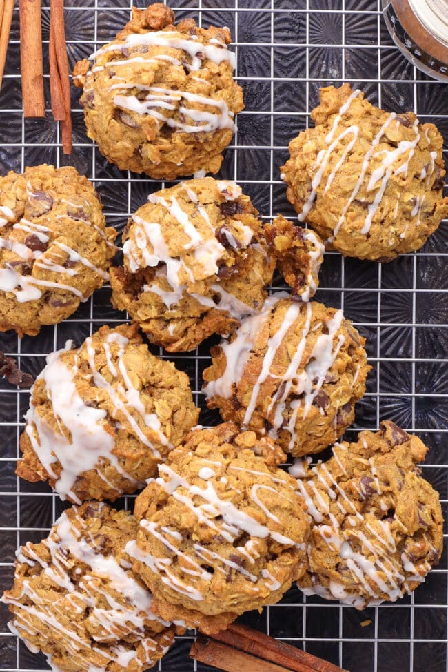 Pumpkin oatmeal chocolate chip cookies drizzled with white icing on a wire cooling rack with cinnamon sticks.