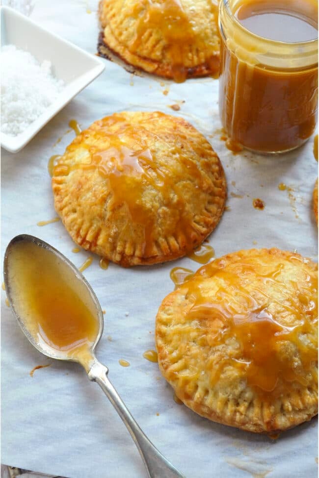 Two hand pies drizzled with caramel sauce, with a small jar of caramel, a spoon, and a dish of coarse salt nearby.